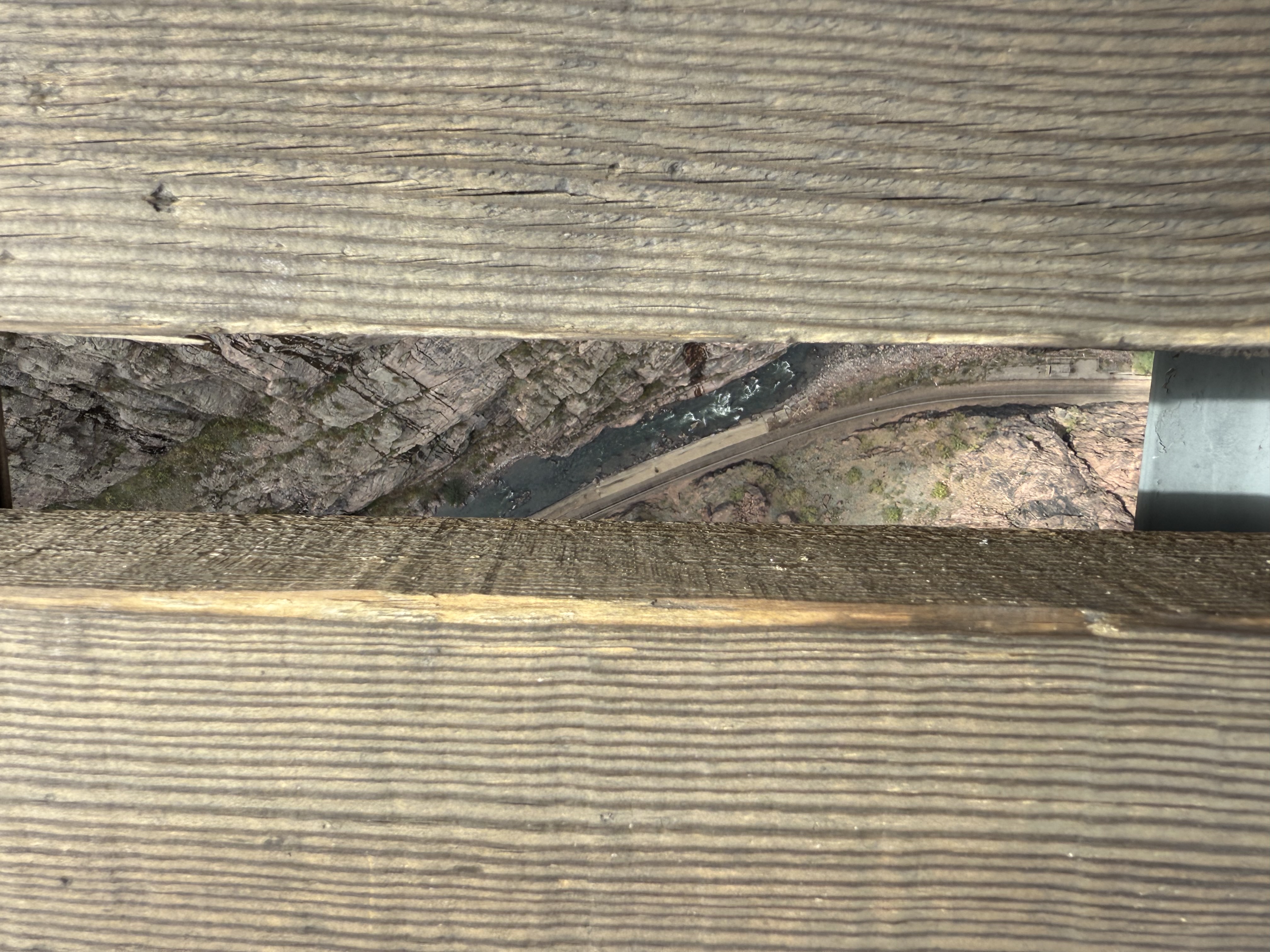 View of a rocky canyon and river far below, seen through a narrow gap between two wide, weathered wooden planks.