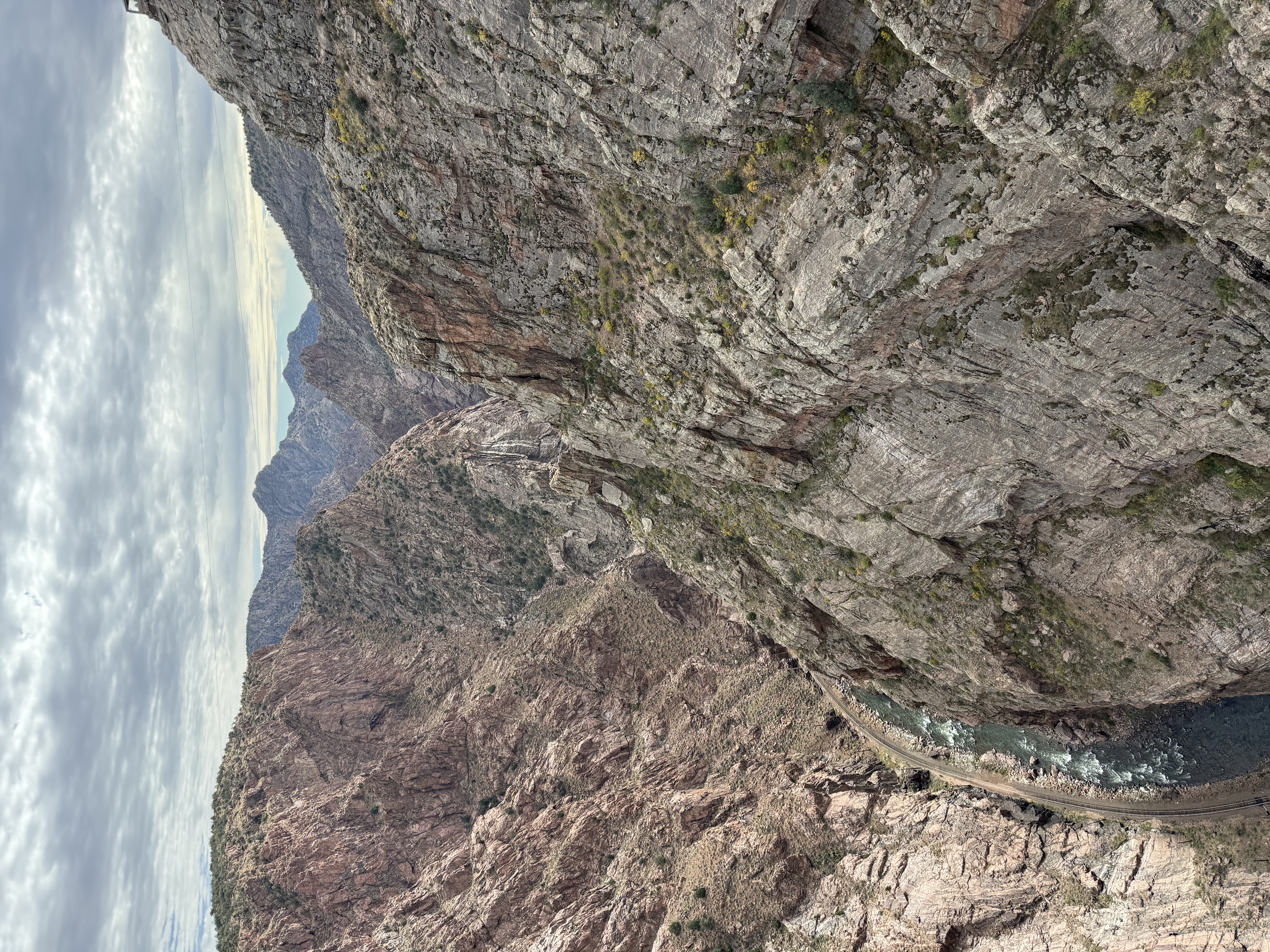 A deep rocky canyon with steep, rugged cliffs and a winding river at the bottom; train tracks run alongside the river. The sky above is cloudy, and distant mountains are visible on the horizon.