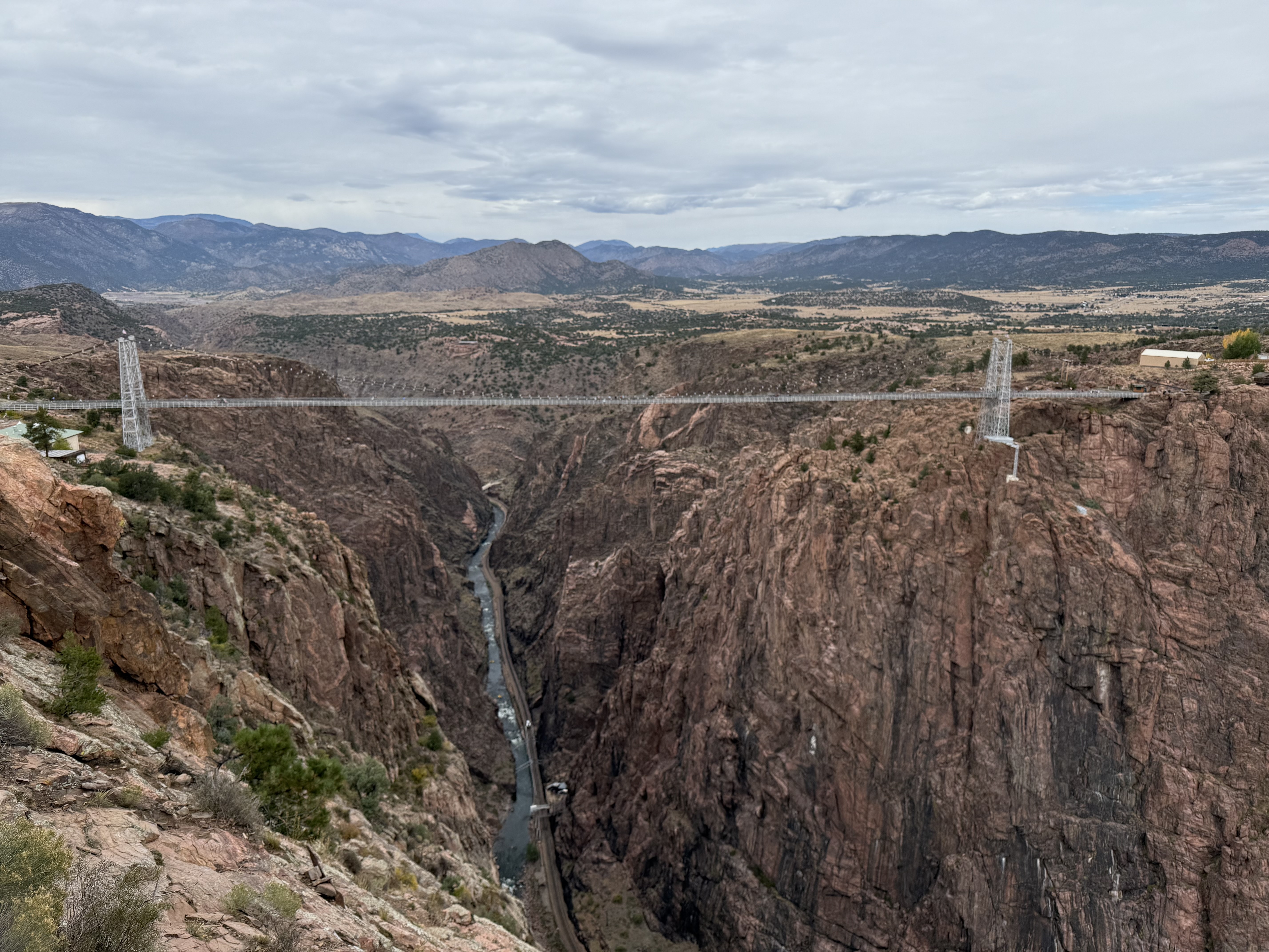 A long suspension bridge spans a deep, rocky canyon with a river at the bottom, surrounded by rugged, reddish cliffs and distant mountains under a cloudy sky.