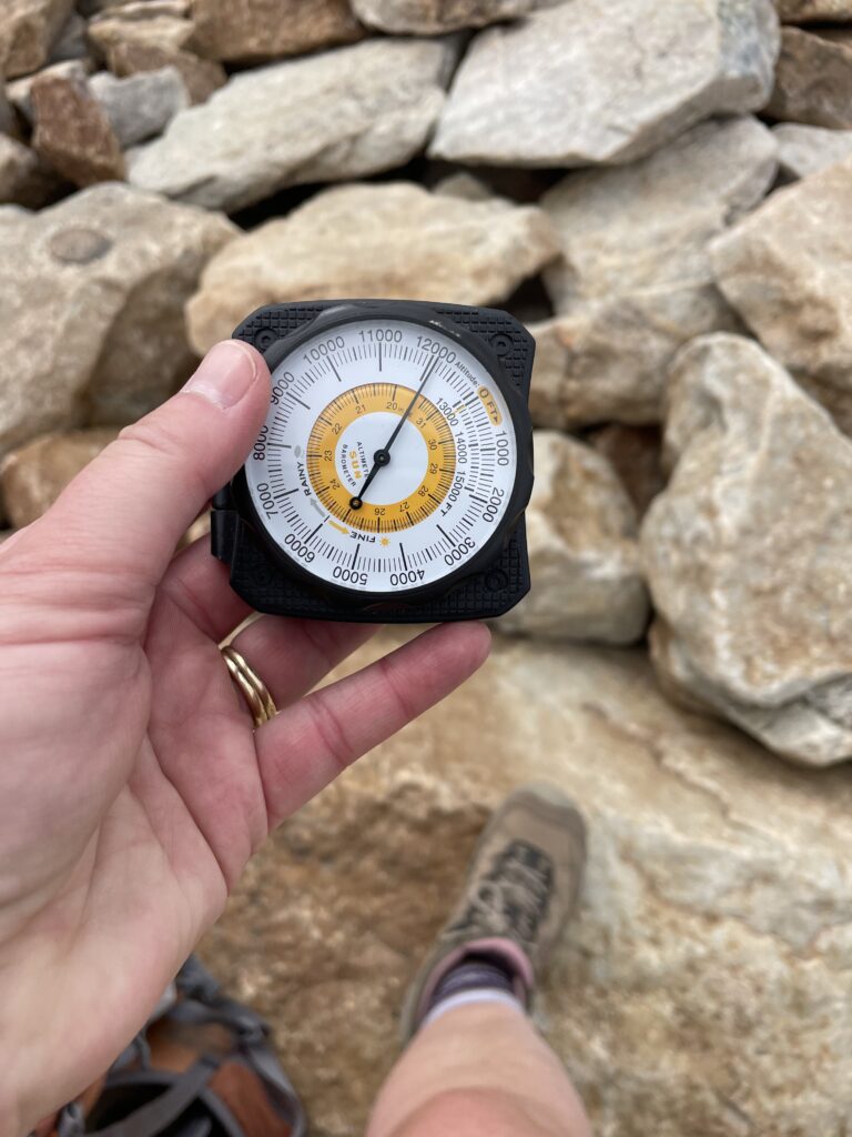 A hand holds an analog altimeter reading about 12,100 feet, with rocky boulders and a hiking boot visible in the background.