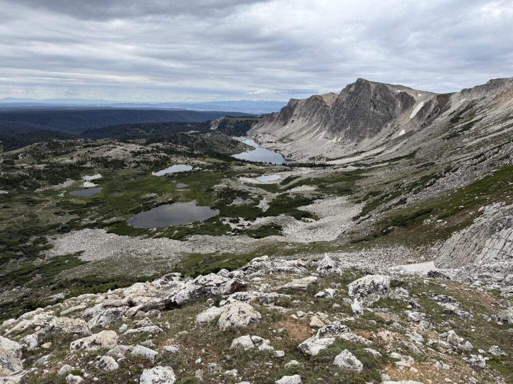 Rocky mountain landscape with several small, alpine lakes and scattered, grassy green, all under a cloudy sky. Rugged peaks rise in the background. The scene is remote and natural.