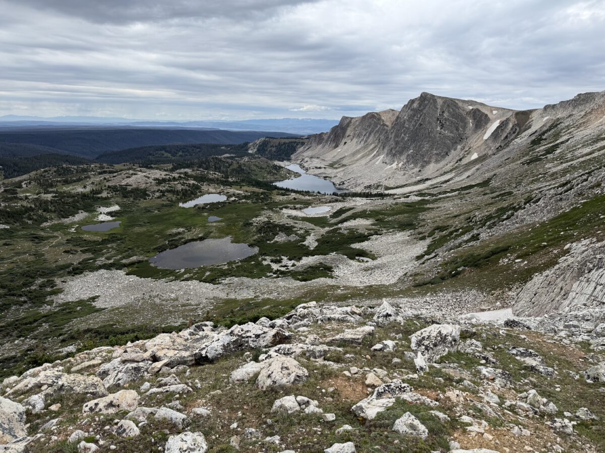 Rocky mountain landscape with several small, alpine lakes and scattered, grassy green, all under a cloudy sky. Rugged peaks rise in the background. The scene is remote and natural.