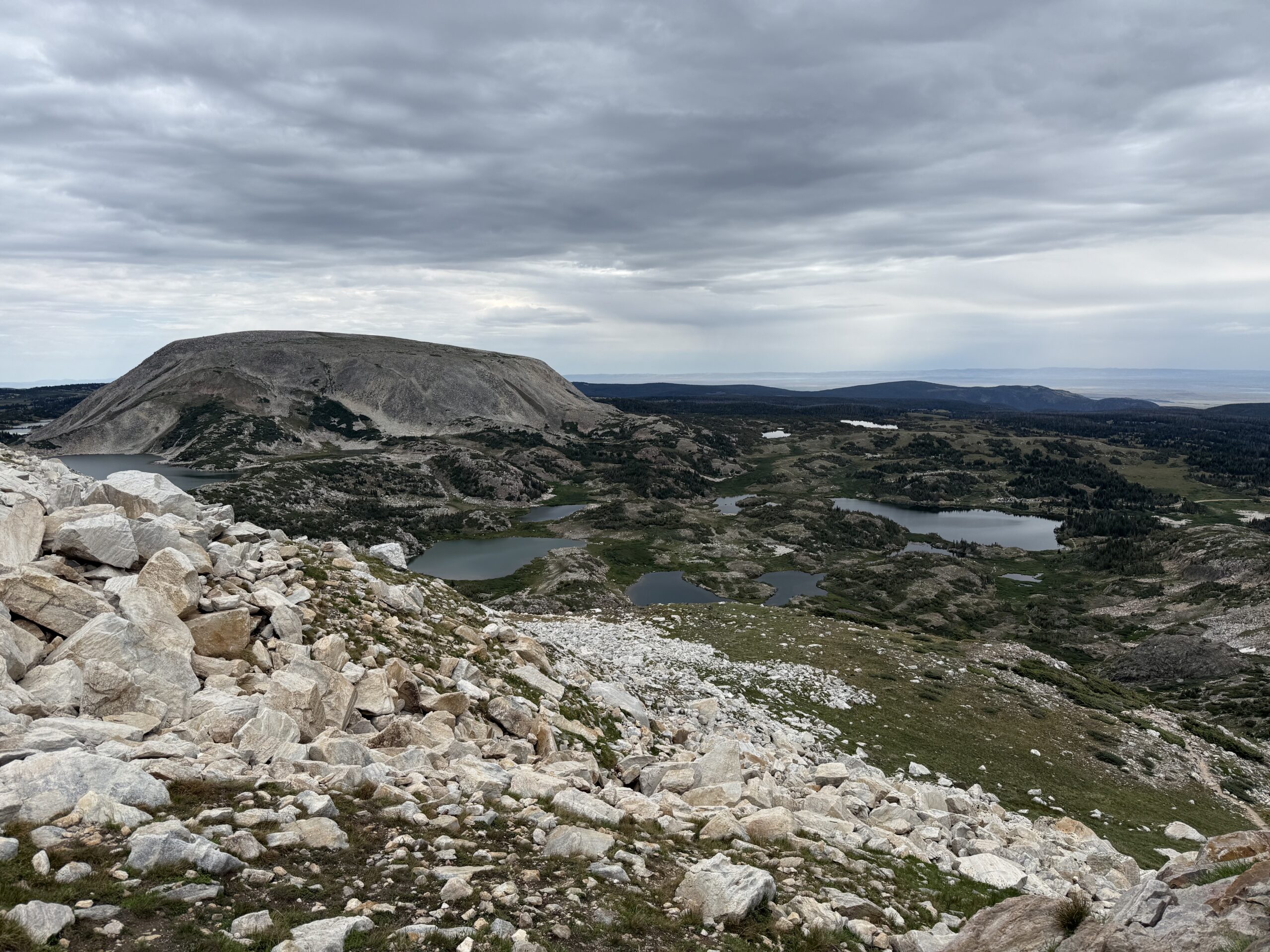 Rocky mountain slope overlooking several small, scattered, apline lakes. A large, rounded mountain sits behind, all under a cloudy sky, with distant forest-covered hills and open plains visible in the background.