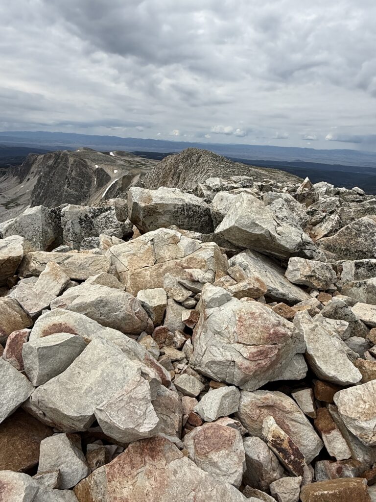 The rocky field on a mountain summit under a cloudy sky, with large boulders in the foreground and distant peaks visible on the horizon. The landscape appears rugged and remote.
